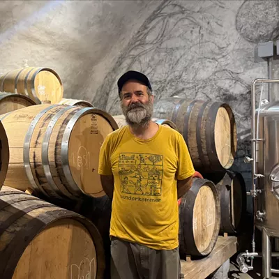 A man wearing a yellow t-shirt stands in front of stacked barrels of beer in a cave
