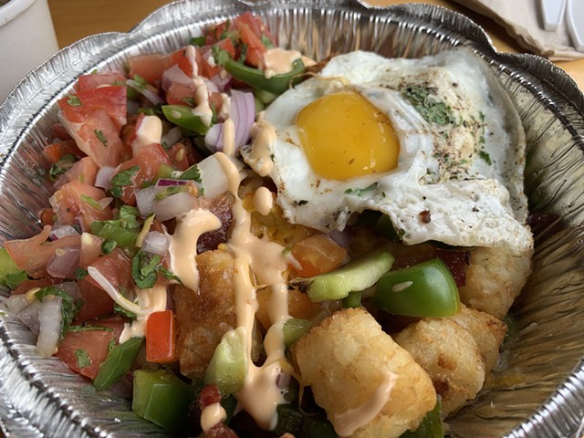 Potatoes covered in vegetables, sauce and a fried egg in a container at wit & grit. in Randolph.