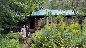 A man and a woman stand in front of a log cabin amidst goldenrod and tall plants