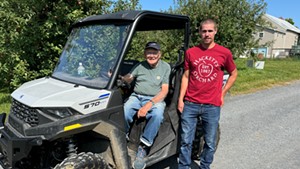 An older man sits in an off-road vehicle, a younger man stands beside him. The younger man's t-shirt says Hackett's Orchard. Behind them is an apple orchard.